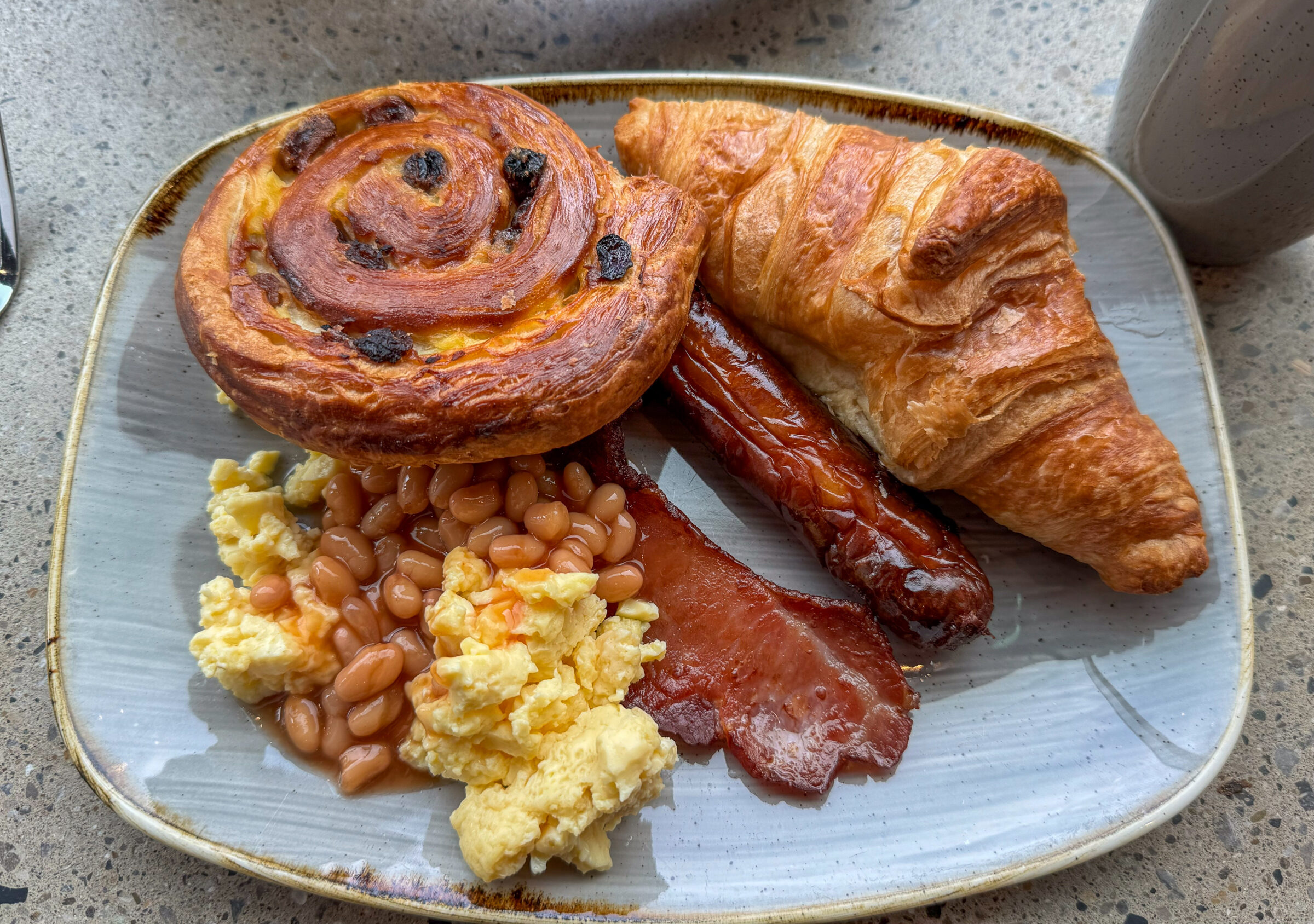 A breakfast plate from a hotel. Two pastries, eggs, beans, bacon, sausage.