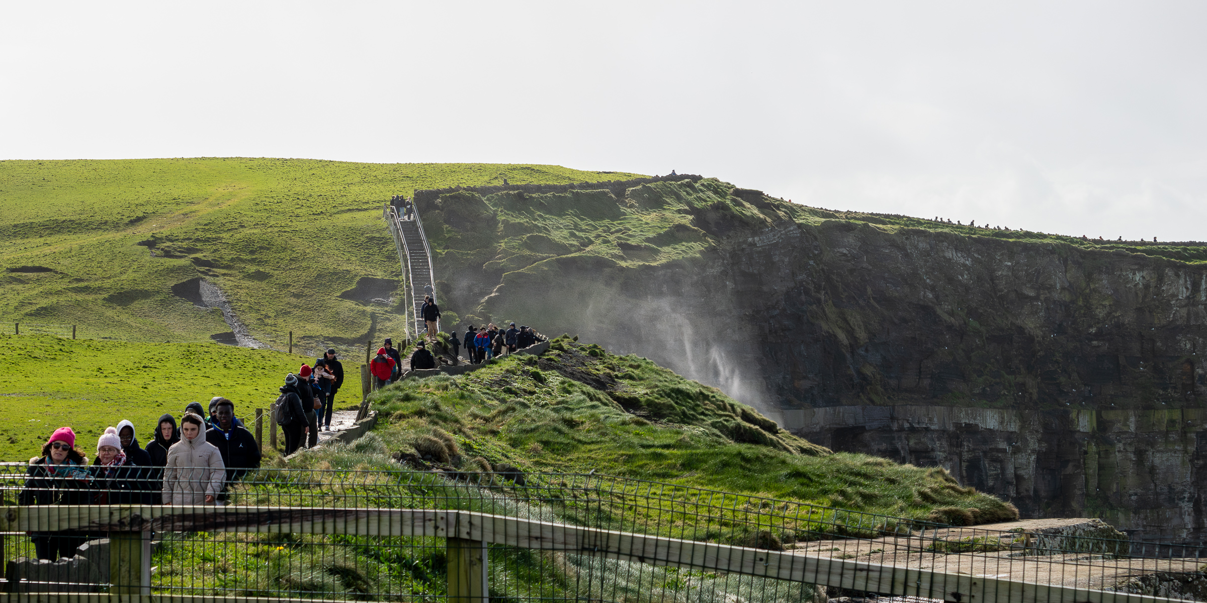 A view of the cliff walk at the Cliffs of Moher. There is green grass along the top of the cliffs. There is ocean spray coming up over the edge from the water below.