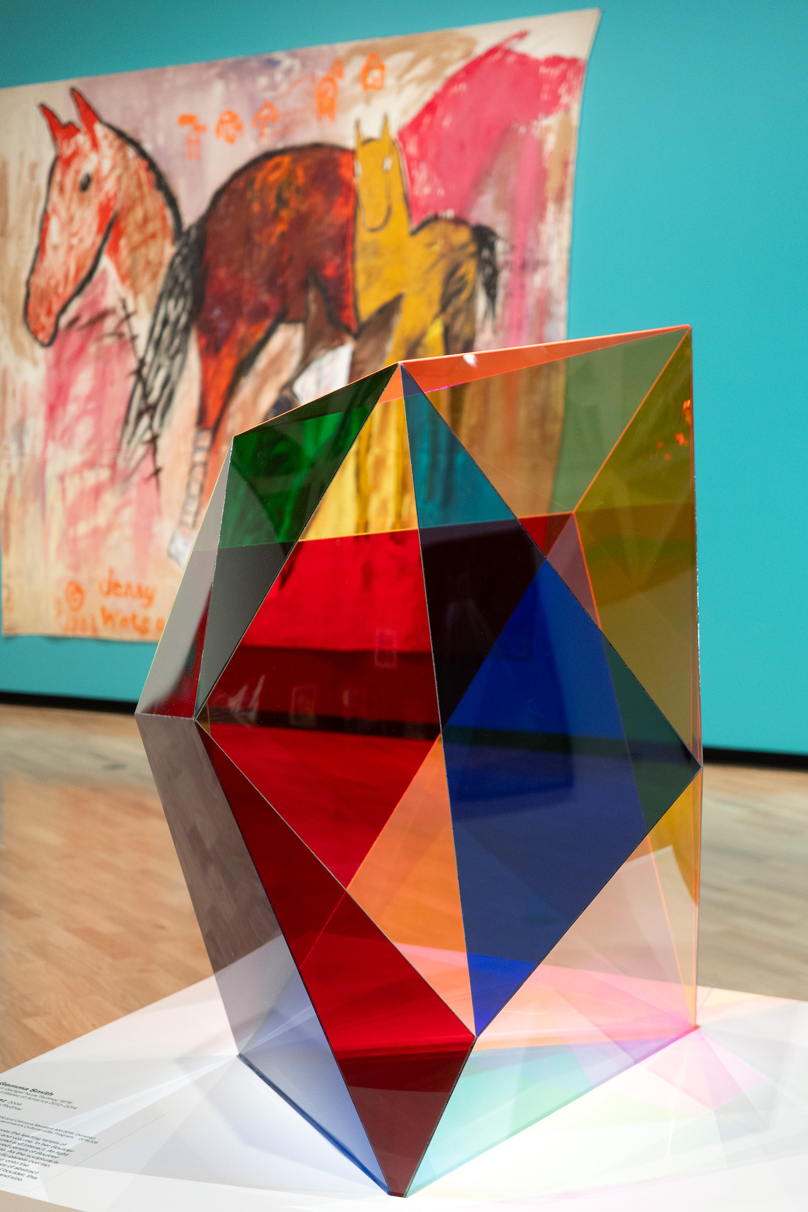A photo of Gemma Smith's Boulder #2 - a colourful semi-transparent sculpture. A large image of a horse in the background on a teal wall.